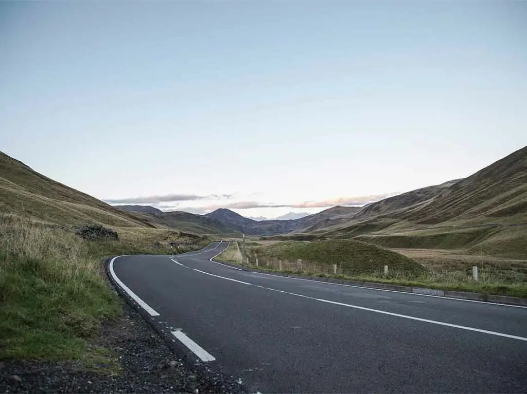 Scenic winding road through Cairngorms National Park in Scotland with rolling green hills and distant mountains under a clear blue sky, ideal for nature tours and sightseeing drives