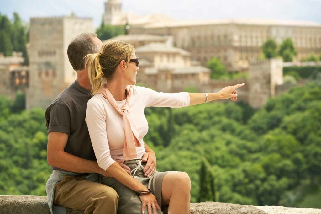 Middle aged couple sitting on wall with woman pointing at view, Granada Spain