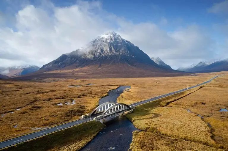 Aerial view of a scenic road and arched bridge crossing a river in Glencoe, Scotland, surrounded by dramatic snow-capped mountains and moorland, highlighting a popular Scottish Highlands travel destination