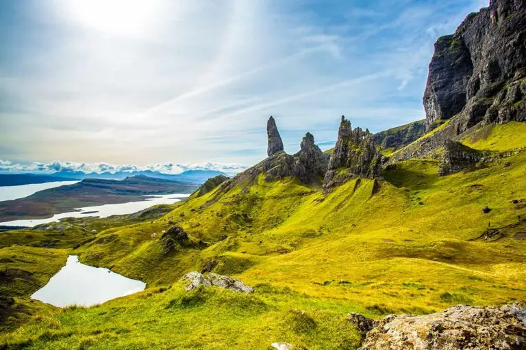 Scenic landscape of the Old Man of Storr rock formations on the Isle of Skye, Scotland, featuring lush green hills, rocky peaks, and distant lochs under a bright blue sky, a popular tourist attraction in Scotland.