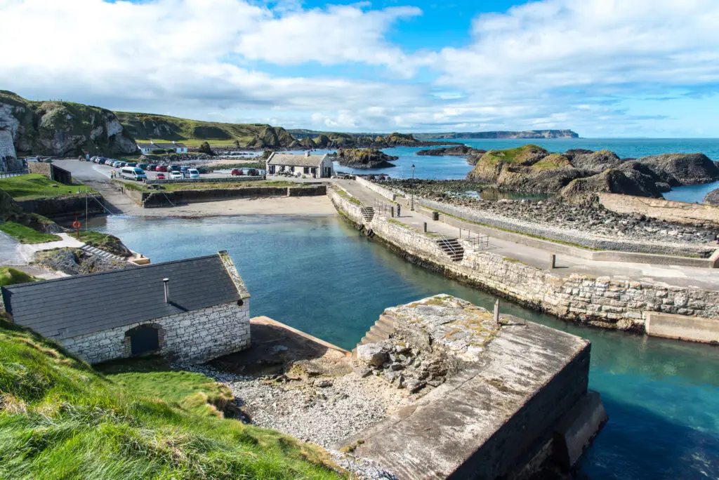 Scenic view of Ballintoy Harbour in Northern Ireland with stone buildings, rocky coastline, clear blue water, and green cliffs under a bright sky