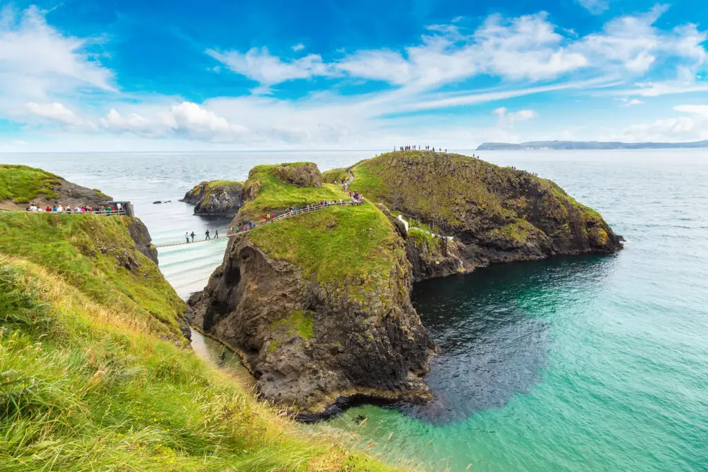 Tourists crossing the Carrick-A-Rede Rope Bridge in Northern Ireland with scenic coastal cliffs, turquoise sea, and lush green landscape under a bright blue sky