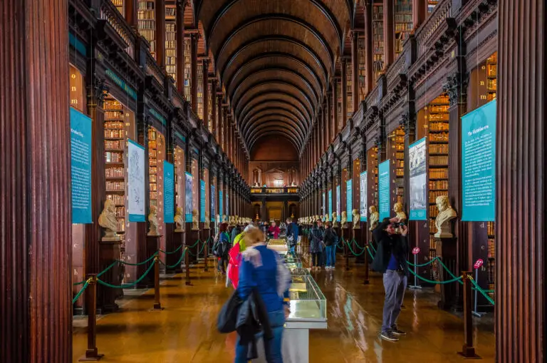 Interior of the Long Room at Trinity College Library in Dublin, Ireland, featuring wooden arches, historic bookshelves, marble busts, and tourists exploring this famous Irish attraction