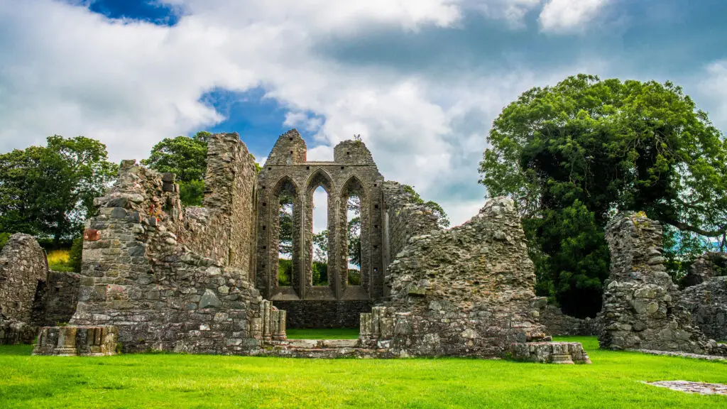Ruins of Inch Abbey surrounded by green grass and trees under a cloudy blue sky in Northern Ireland, a popular historic site for Ireland tours and sightseeing