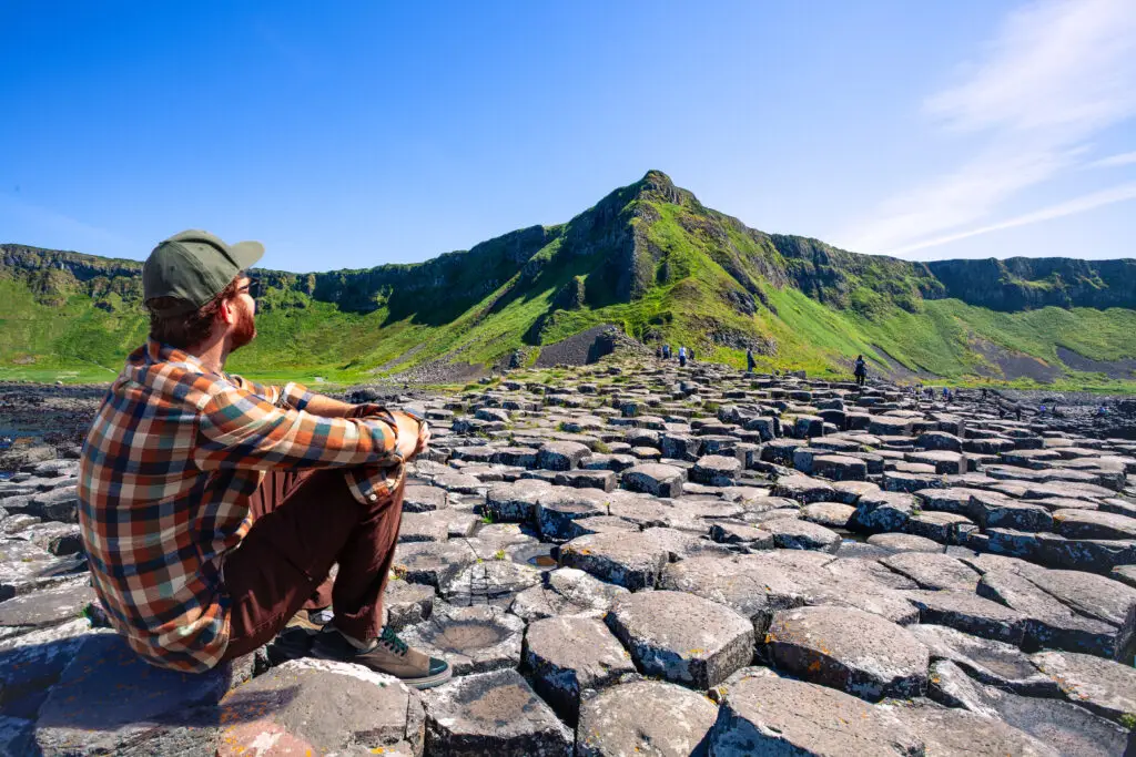 Tourist sitting on basalt columns at the Giant’s Causeway in Northern Ireland with green cliffs and blue sky in the background
