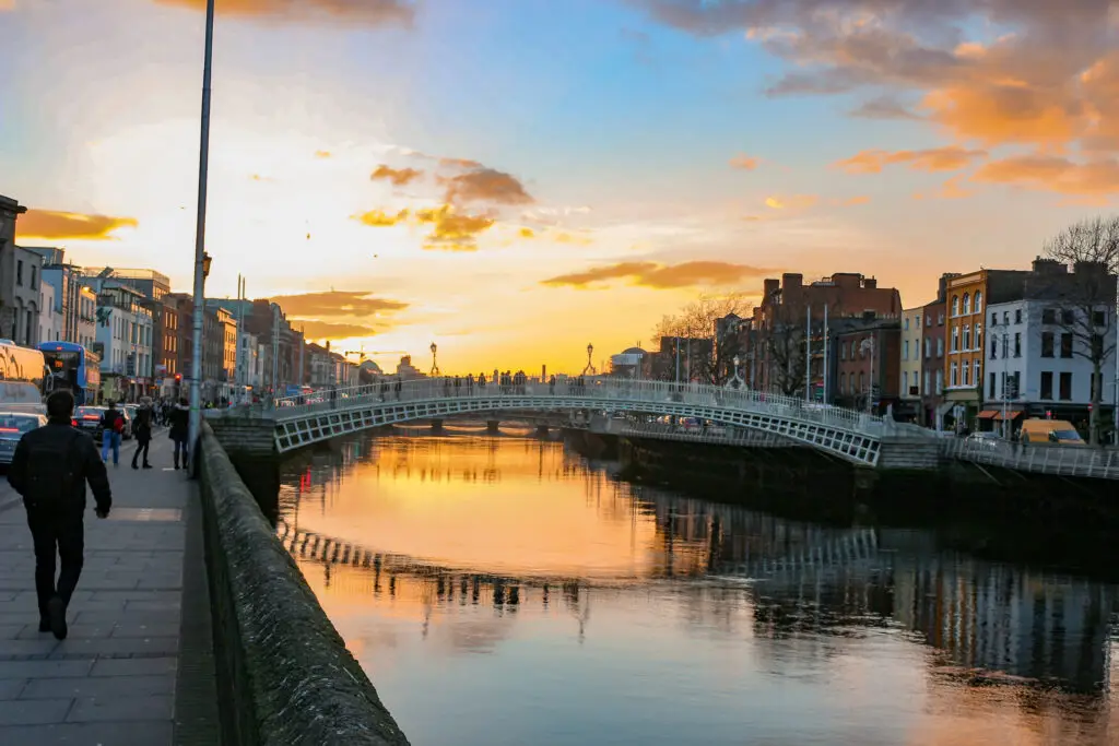 Ha’penny Bridge over the River Liffey at sunset in Dublin, Ireland with people walking along the riverside and reflections of city buildings in the water