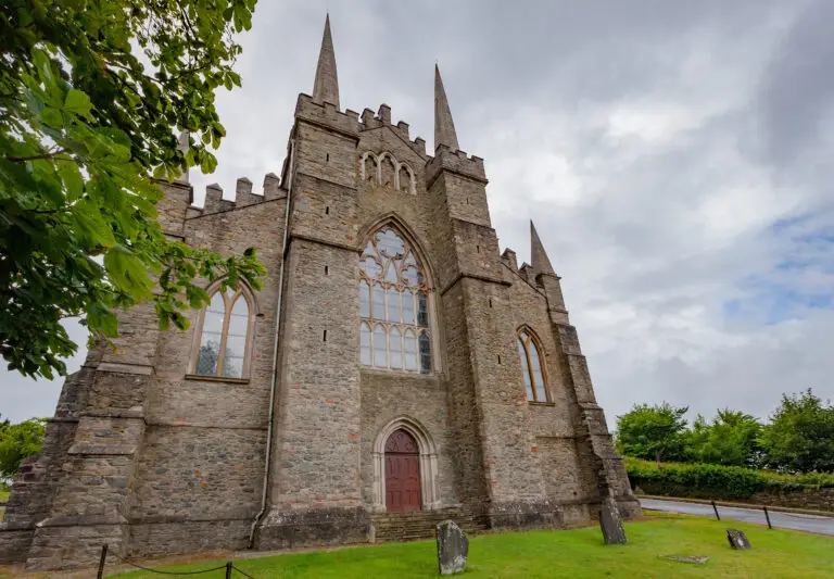 Historic Down Cathedral in Downpatrick, Northern Ireland, featuring Gothic architecture with stone walls and pointed spires surrounded by greenery under a cloudy sky