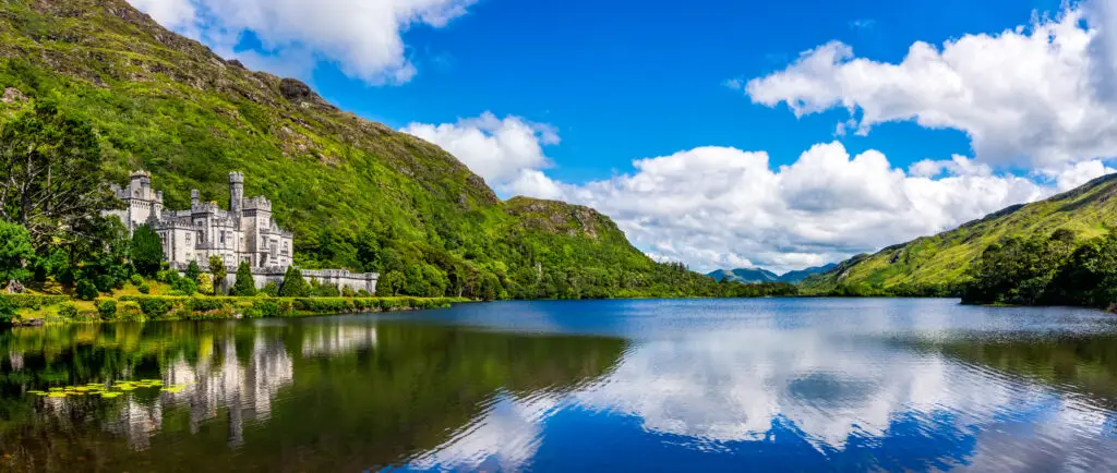 Kylemore Abbey reflected in Pollacapall Lough surrounded by lush green hills under a bright blue sky in Connemara, County Galway, Ireland