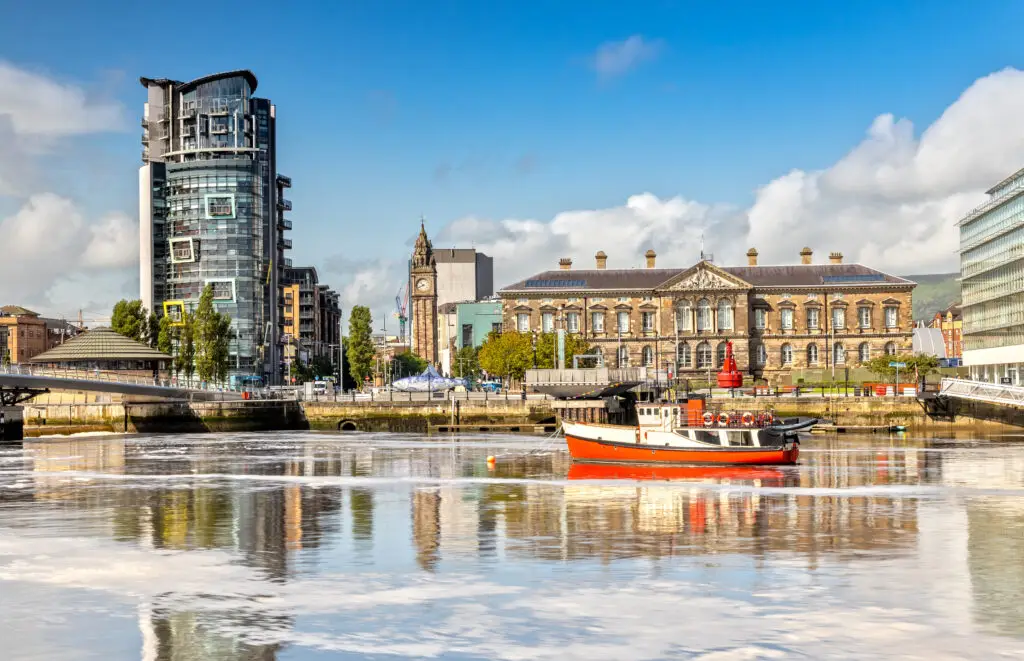 The Custom House and Lagan River waterfront with a red boat and modern city buildings in Belfast, Northern Ireland, on a sunny day