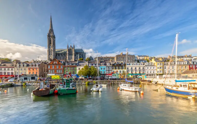 Colorful waterfront buildings and boats in the harbor of Cobh, Ireland with St. Colman’s Cathedral in the background on a sunny day