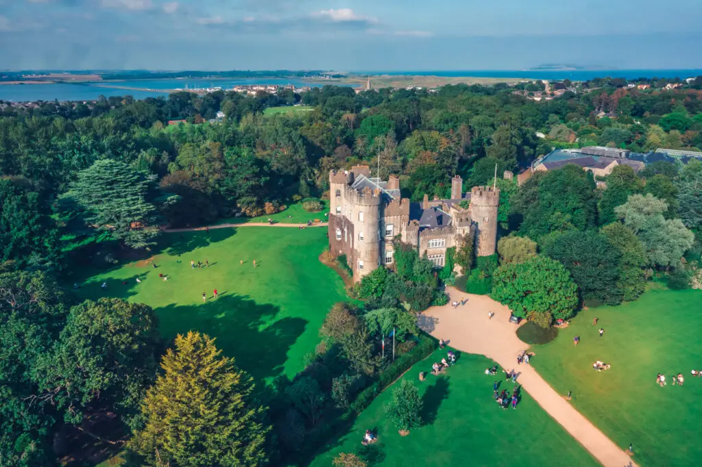 Aerial drone view of Malahide Castle in Dublin, Ireland, surrounded by lush green gardens and visitors enjoying the scenic historic landmark and coastal landscape.