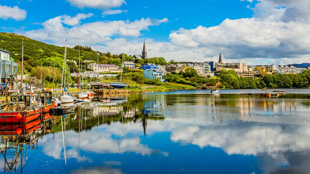 Colorful waterfront view of Clifden, Ireland with sailboats moored along the harbor, reflections on the calm water, and vibrant houses set against green hills under a bright blue sky