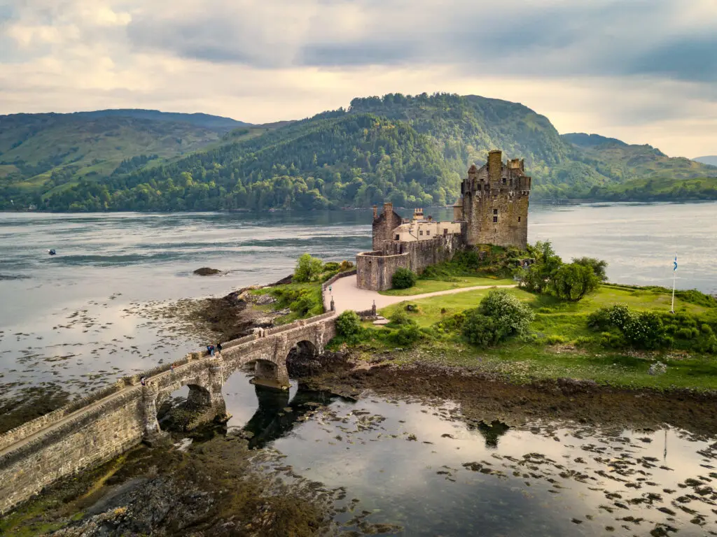 Eilean Donan Castle in Scotland with stone bridge over water, surrounded by lush green hills and scenic landscape popular with tourists