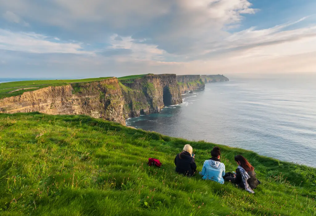 Tourists sitting on green cliffs overlooking the Atlantic Ocean at the Cliffs of Moher, a famous coastal attraction in County Clare, Ireland