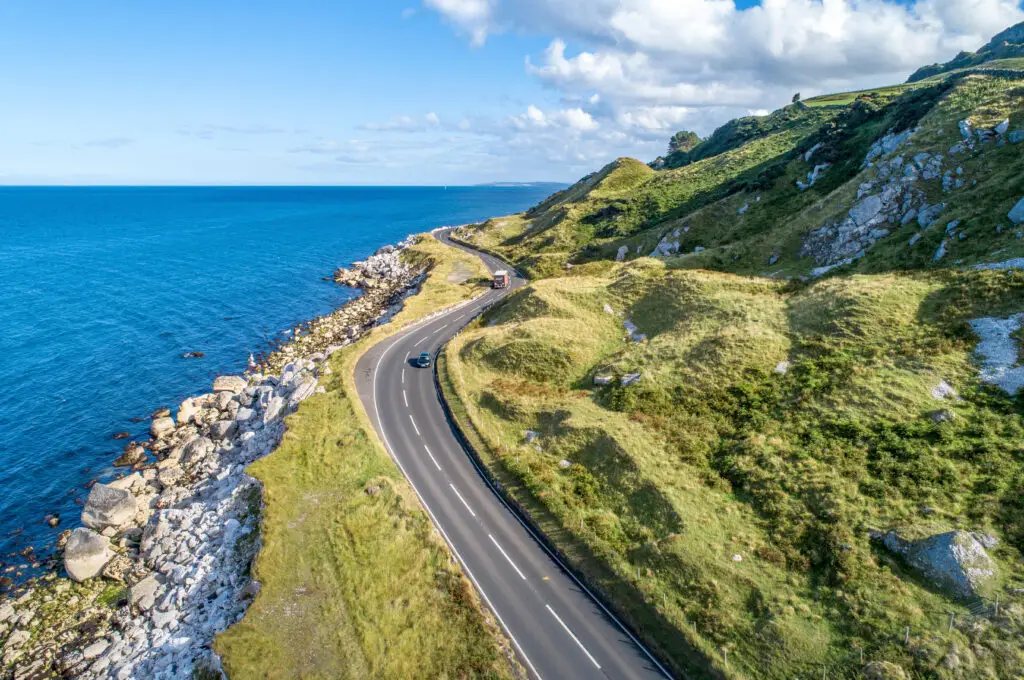 Scenic view of the Antrim Coastal Road in Northern Ireland with winding roadway, green cliffs, and blue sea under a bright sky