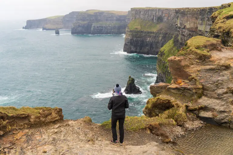 Tourist standing on cliff edge overlooking the dramatic Cliffs of Moher and Atlantic Ocean in County Clare, Ireland