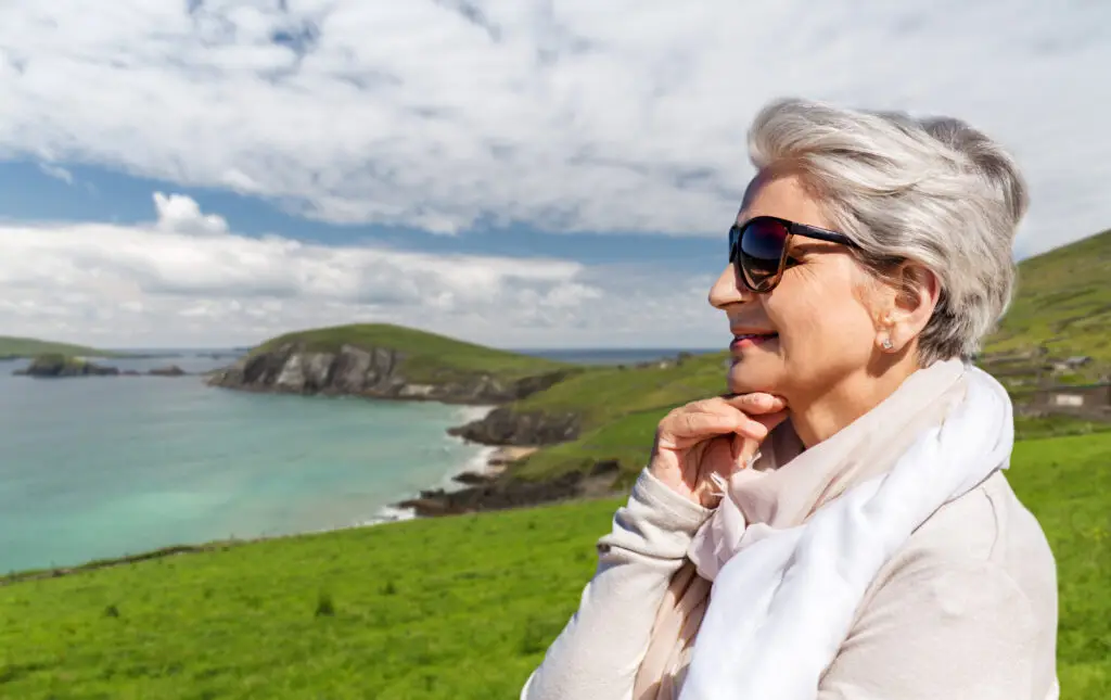 Smiling woman enjoying scenic coastal view of the Dingle Peninsula, Ireland, with cliffs, turquoise water, and green hills in the background on a clear day