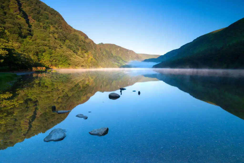 Scenic view of Glendalough Park in Ireland with calm lake reflections, surrounding green mountains, and morning mist creating a peaceful landscape ideal for nature photography and tourism tours