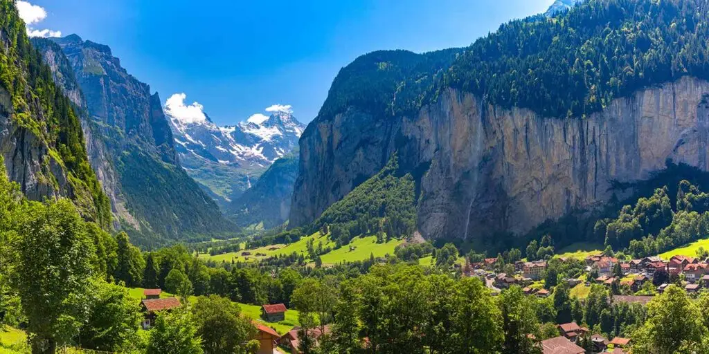 Scenic panoramic view of Lauterbrunnen Valley in Switzerland featuring alpine mountains, towering cliffs, waterfalls, and charming village houses surrounded by lush green meadows under a bright blue sky